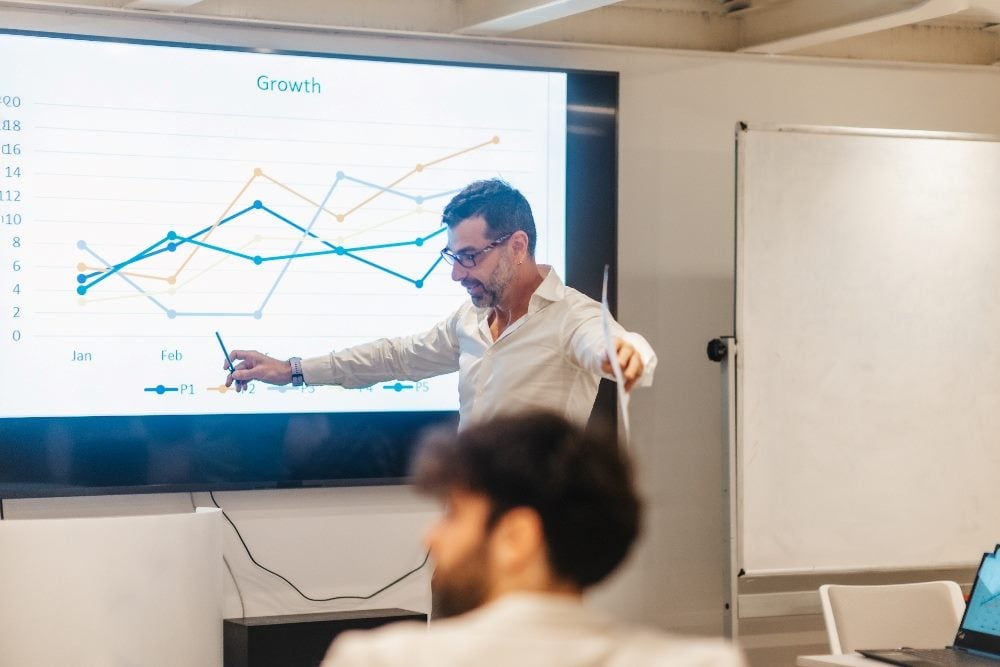 Un hombre de negocios con gafas y lápiz y papel analiza un informe de crecimiento proyectado en una pantalla detrás de él en un segmento sobre la toma de decisiones basada en datos.