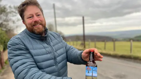 La imagen tiene una botella de sidra Tom Bull. El fuftor lleva una chaqueta azul y está parada al lado de una carretera. La foto fue tomada en un día nublado.