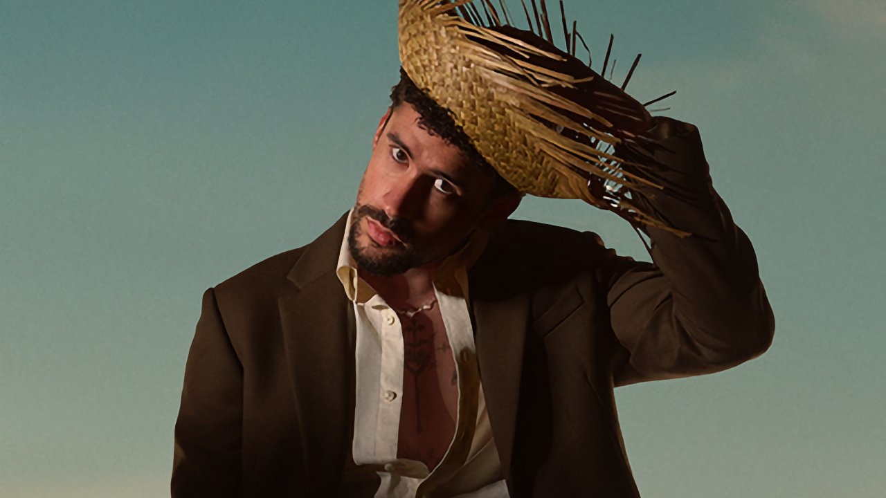 Una persona que lleva una chaqueta y una camisa oscuras abiertas ajusta un sombrero de paja contra un fondo de cielo despejado.