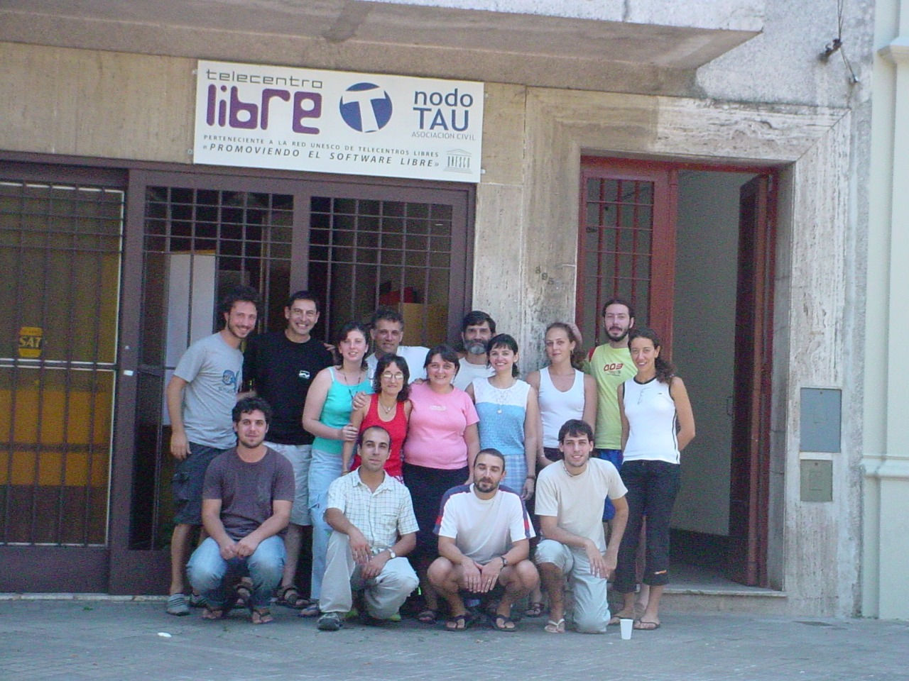 Grupo de personas posando frente a una casa con ventanas cerradas y puerta abierta. Un letrero encima de la puerta y la ventana anuncia Telecentro Libre Nodo TAU.