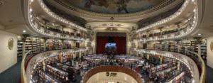 Una vista de cámara de personas caminando entre estantes de metal y madera llenos de libros sobre un suelo alfombrado. Múltiples niveles rodean el espacio abierto principal con más estanterías, iluminación decorativa y tallas arquitectónicas intrincadas. Al fondo hay un alto telón rojo y el escenario.