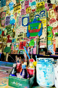 Botellas de refresco y pinceles cubiertos de pintura se encuentran sobre una mesa frente a un gran mural cubierto de rostros brillantes, robots y carteles.